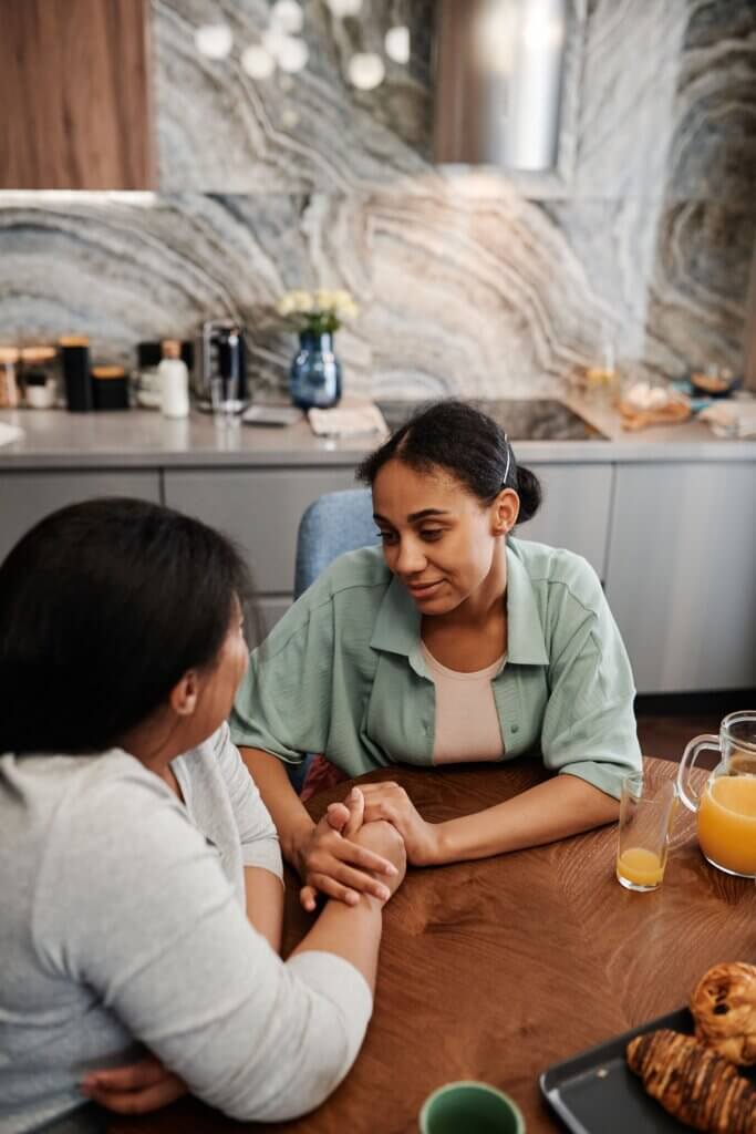 Dos mujeres tomadas de las manos en una conversación cercana y de apoyo en una mesa de cocina, imagen que ilustra el acompañamiento a un adolescente en dificultad