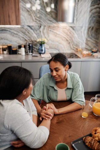 Dos mujeres tomadas de las manos en una conversación cercana y de apoyo en una mesa de cocina, imagen que ilustra el acompañamiento a un adolescente en dificultad